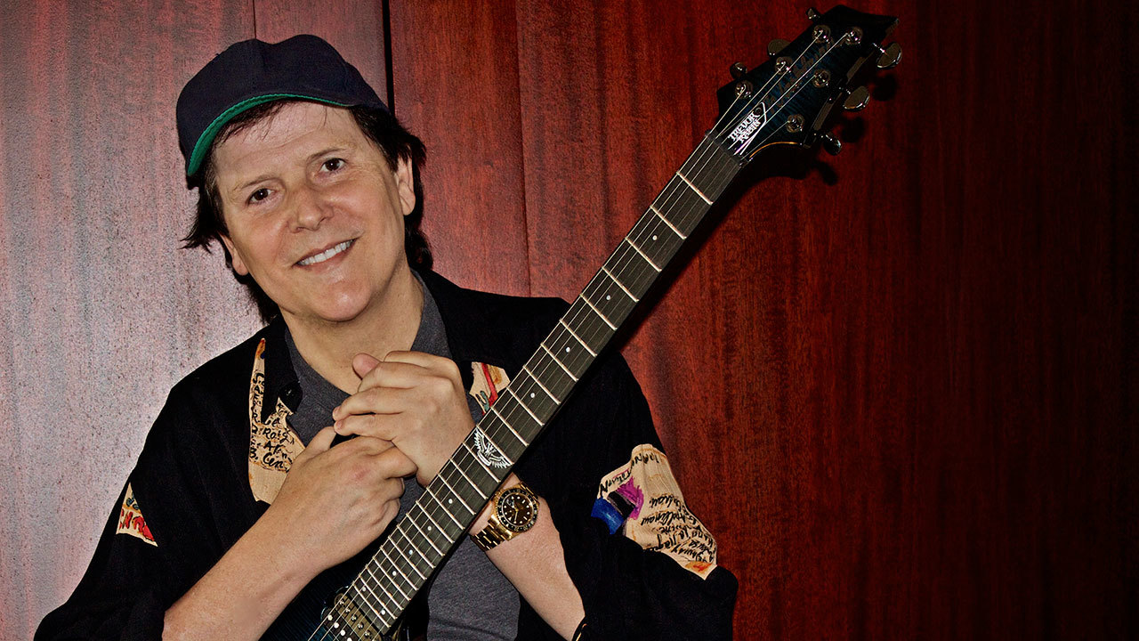 A photograph of Trevor Rabin holding a guitar and smiling