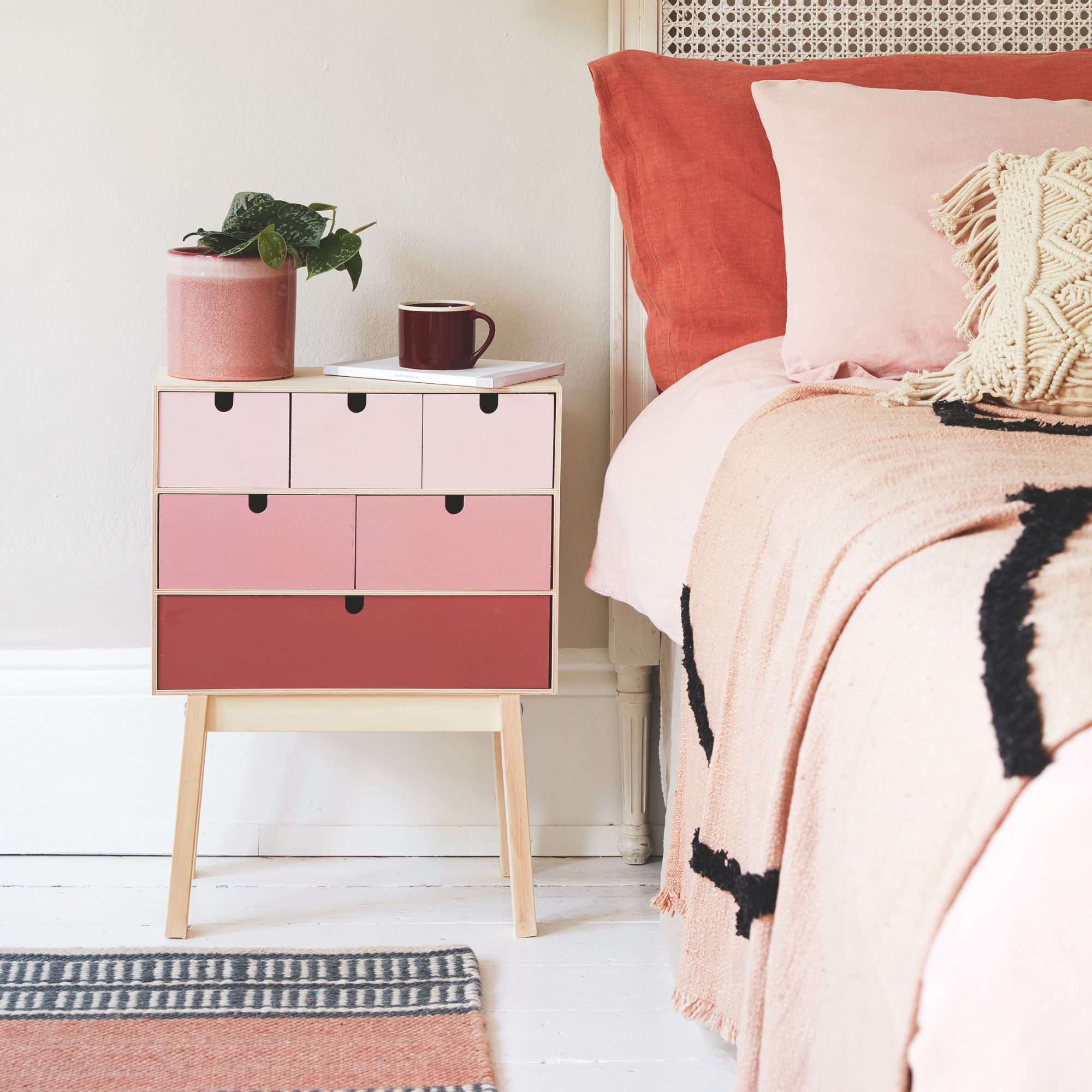 Dormitorio pintado blanco con una mesita de noche pintada rosa y una maceta rosa encima de ella, al lado de una cama con ropa de cama rosa