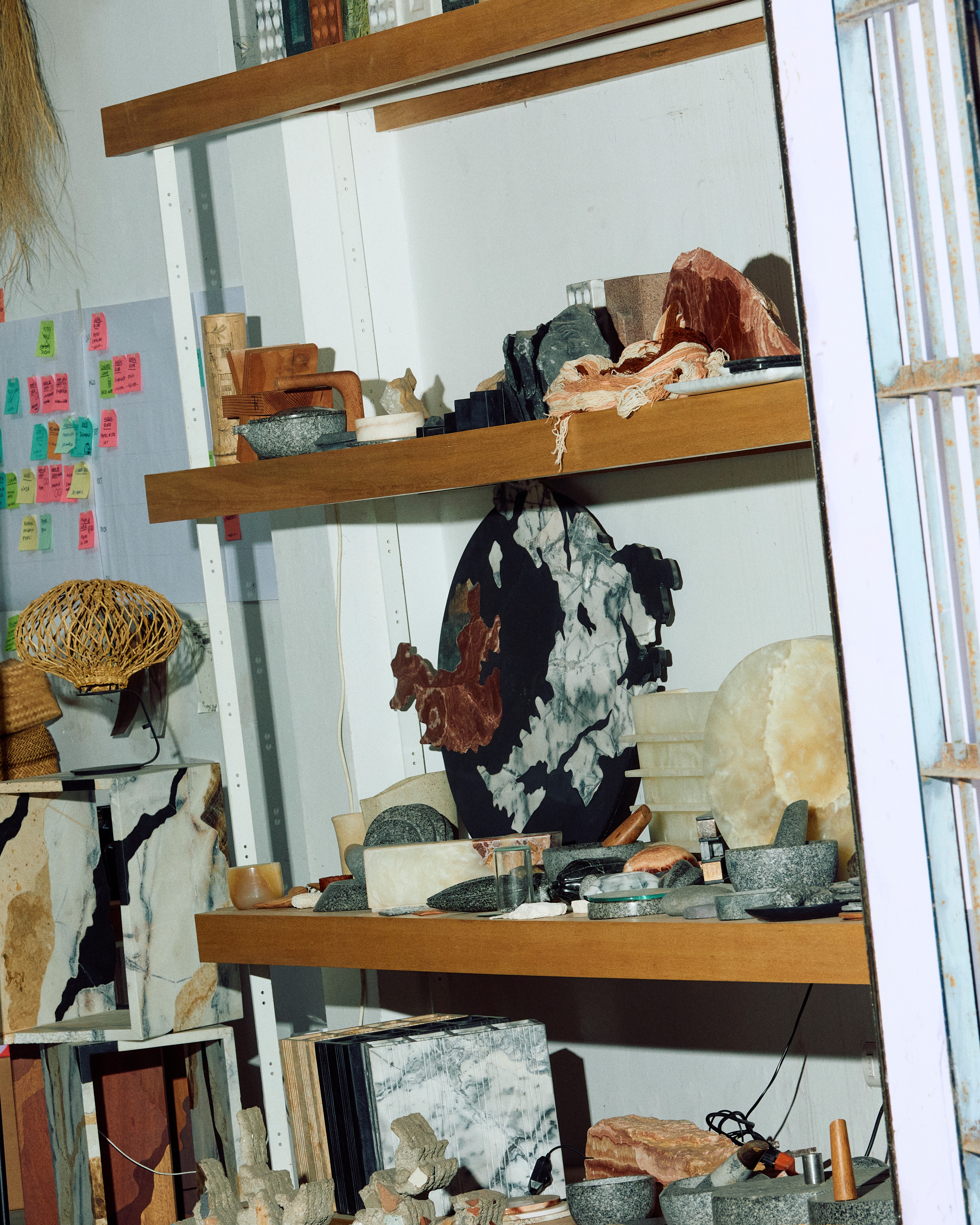 Interiors of an artisan workshop filled with rattan, wood, stone, and ceramic sculptures and light, among which sit or stand its artisans.