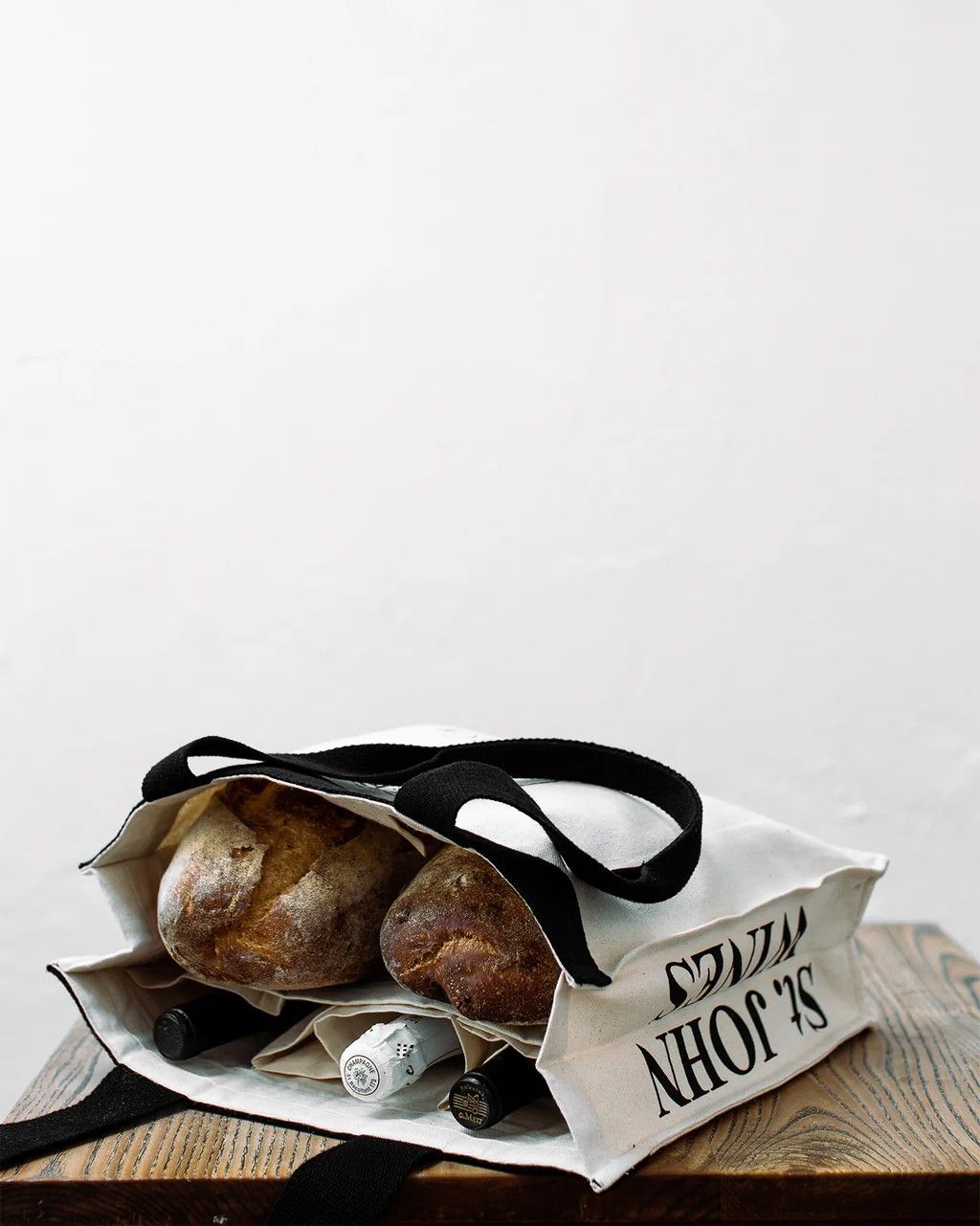 A black and white tote bag with the sketch of a person sniffing wine and the writing "SNIFFING" is being hold by a person entirely dressed in black in an industrial, naturally lit space, and is seen resting while filled with bread and wine on a wooden table in the image next to the previous.