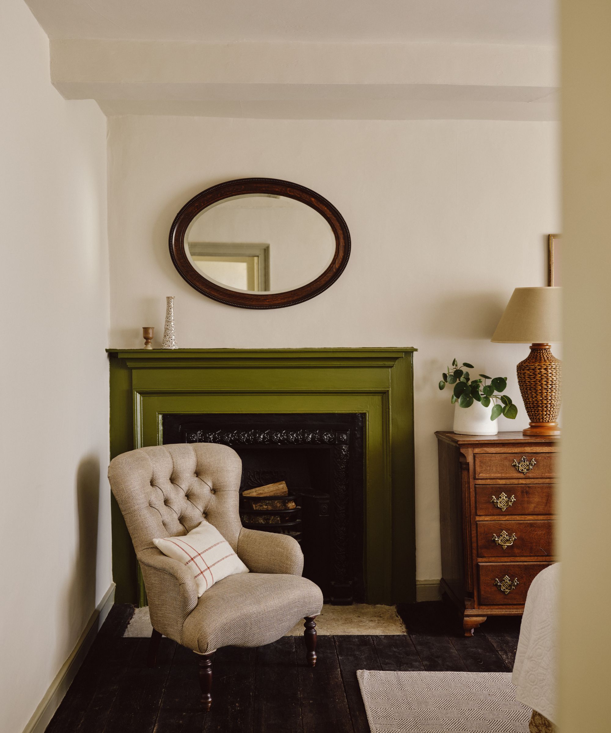 A farmhouse bedroom with white walls, a moss green fireplace, an oval mirror, and a warm gray armchair.