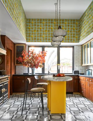 Kitchen with yellow sculptural island, 70s patterned floor tiles and wallpaper, pendant lights and bar stools