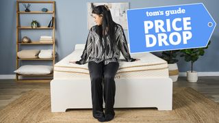 A woman sits on the foot of a Saatva Classic Mattress in a bedroom, a Tom's Guide price drop deals graphic (right)