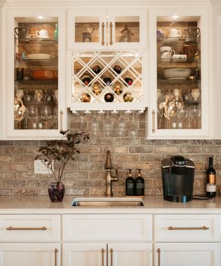 A kitchen counter with a purple vase of branches, a brushed gold sink and faucet, a black coffee maker, and a bottle of wine, with a white kitchen cabinet above it filled with jars, glassware, and bottles of wine and white cabinets with gold handles beneath it