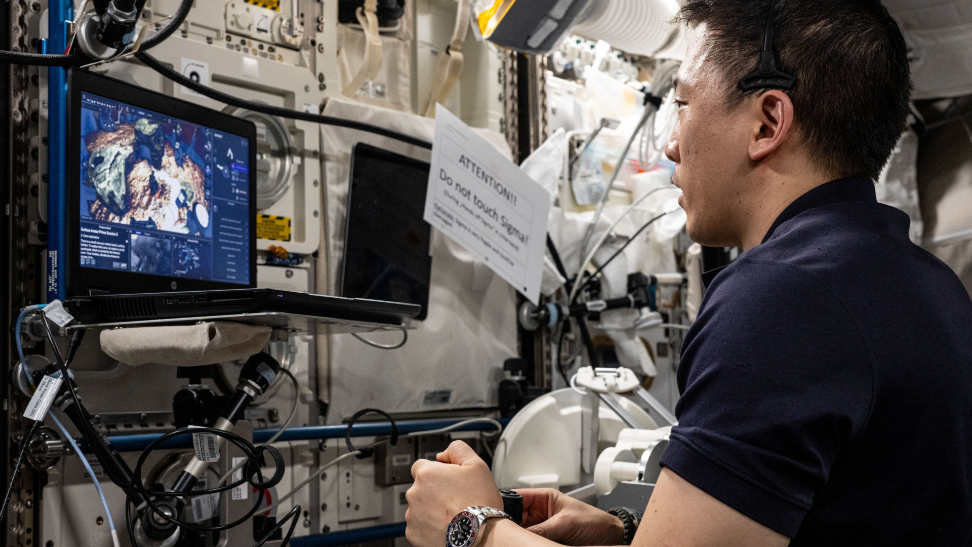 A man wearing a dark blue shirt and metal watch  uses a controller while watching a screen