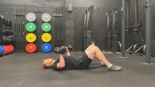 FItness writer Harry Bullmore performs a dumbbell floor press in a gym. He lies on the floor with his knees bent and back flat. His elbows are also bent and close to his sides, as he holds a pair of dumbbells in his hands, just above chest height. Behind him we see colorful weight plates and gym equipment.