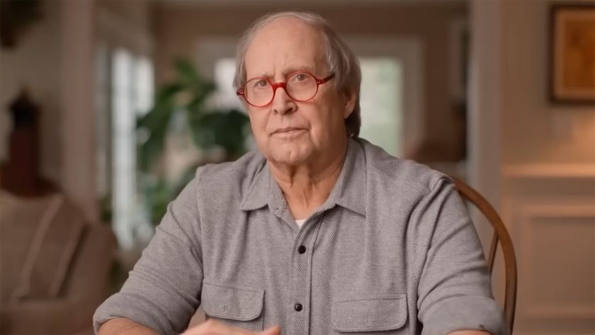 Chevy Chase sits with his hands folded and red glasses on at a dining room table.