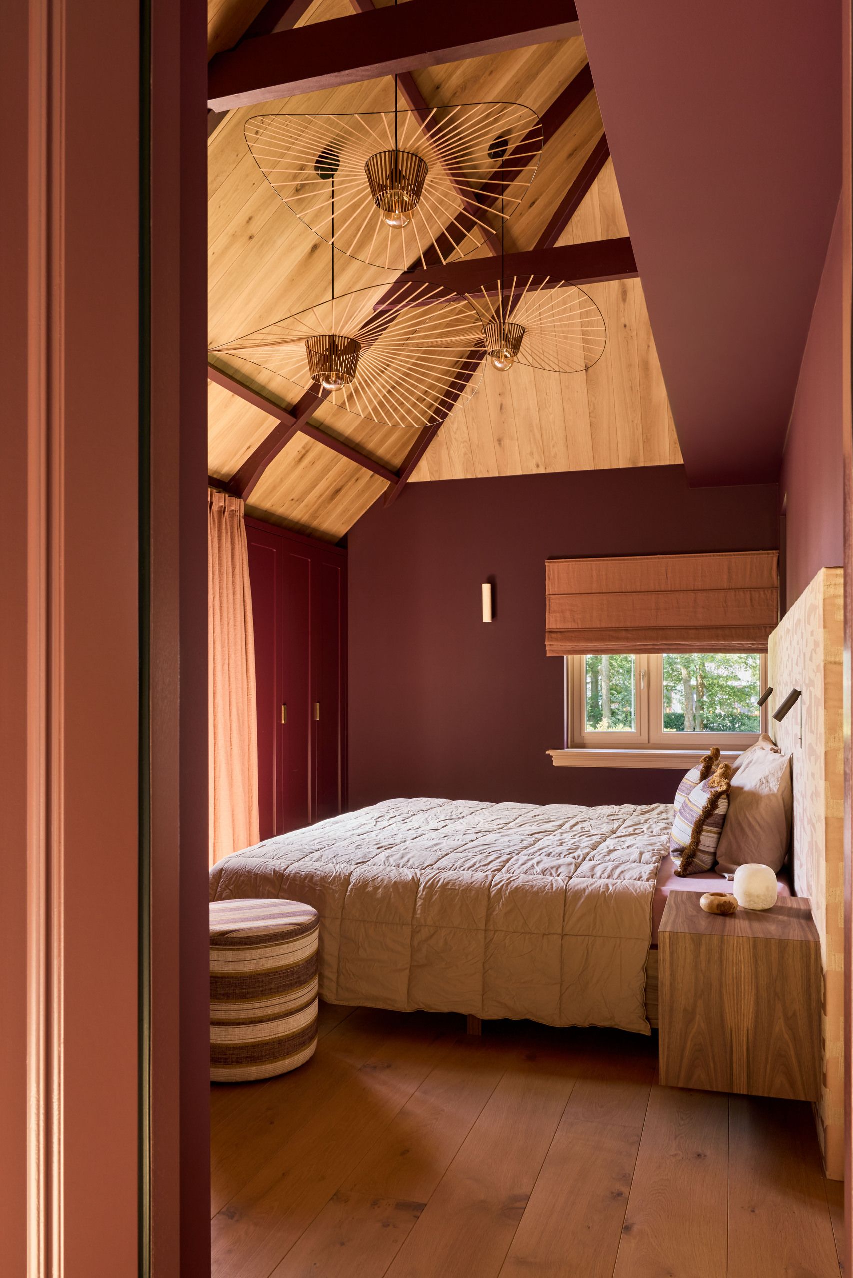 Image of a dusty burgundy bedroom with high ceilings, off-white bedding, and three sculptural pendant lights in the center of the room.