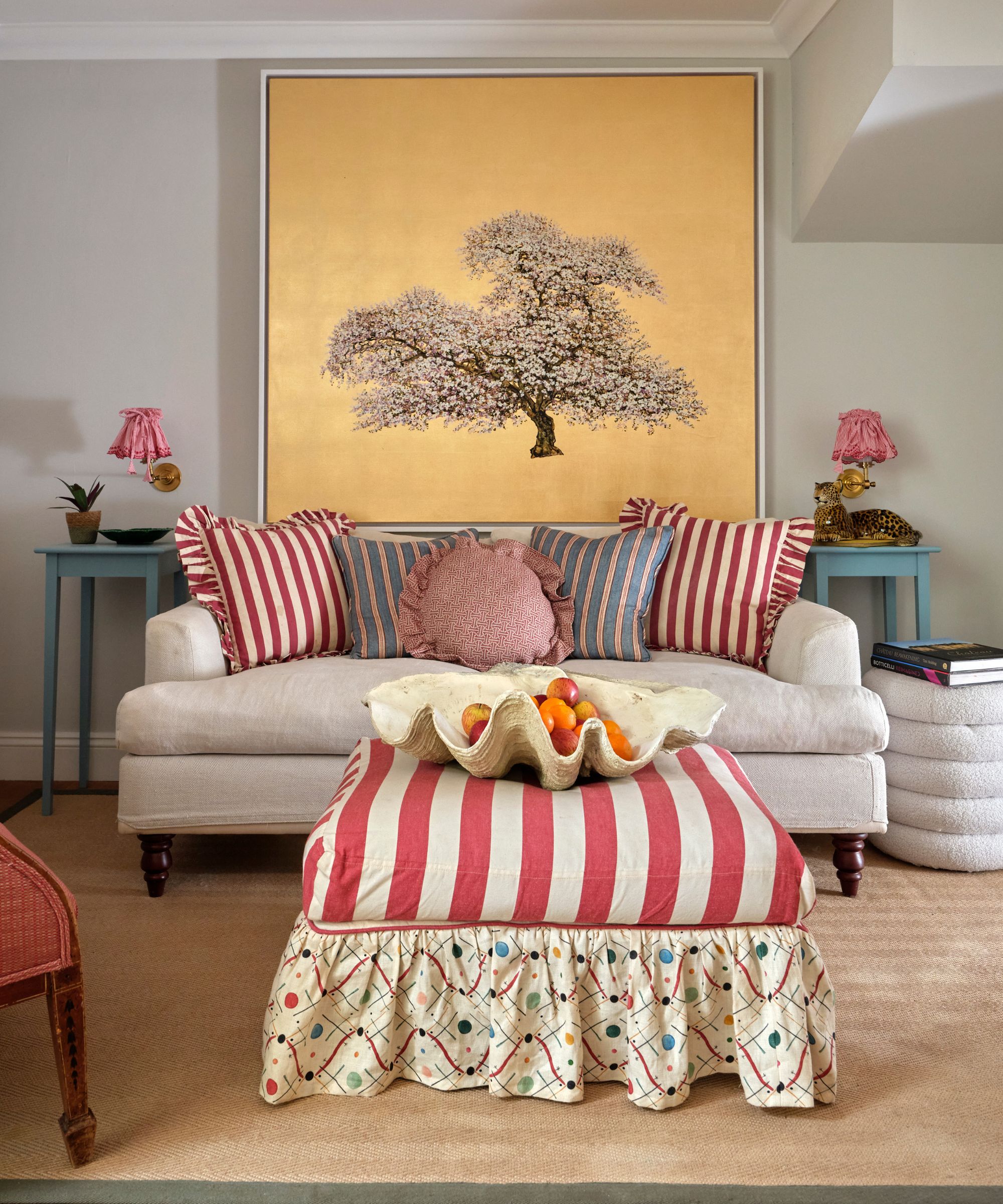 A cozy living room featuring a white sofa with red striped cushions and a large gold-leaf painting of a cherry blossom tree. A red and white striped ottoman sits in the foreground on a sisal rug.