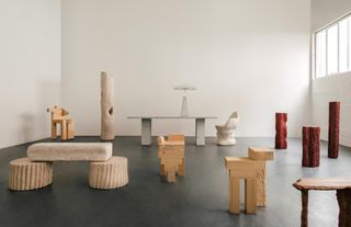 In an industrial gallery space, a collection of organically hued furniture pieces including coffee tables, chairs, columns, benches, and tables are scattered across the room.