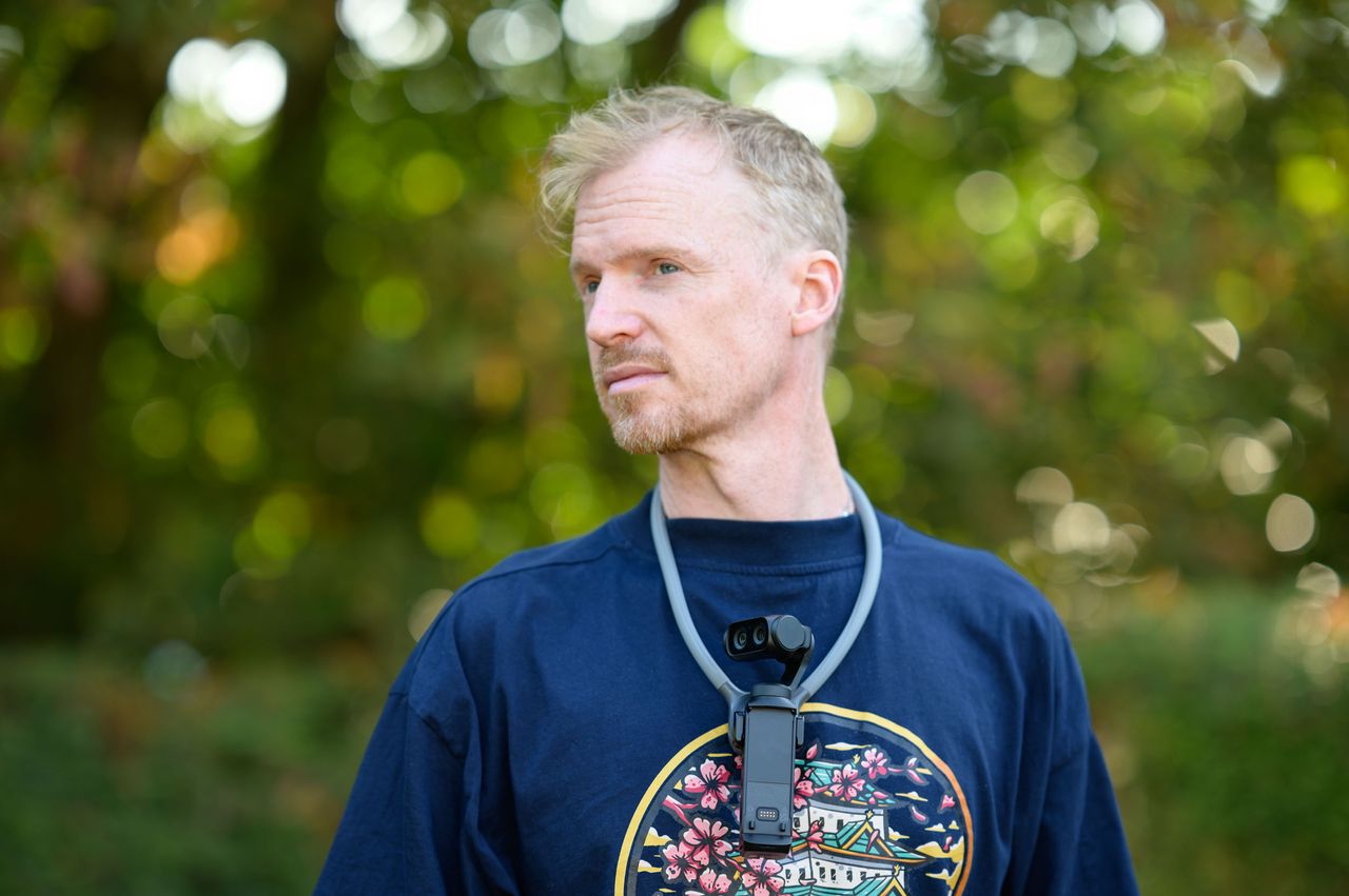 Tim Coleman wearing the Insta360 POV Head Tracker accessory in his ear, and Luna Ultra camera on his chest attached to a neck mount, outdoors with dappled light in background foliage