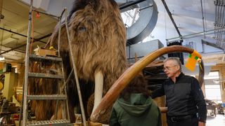 Two people stand next to the model of a woolly mammoth in a workshop.