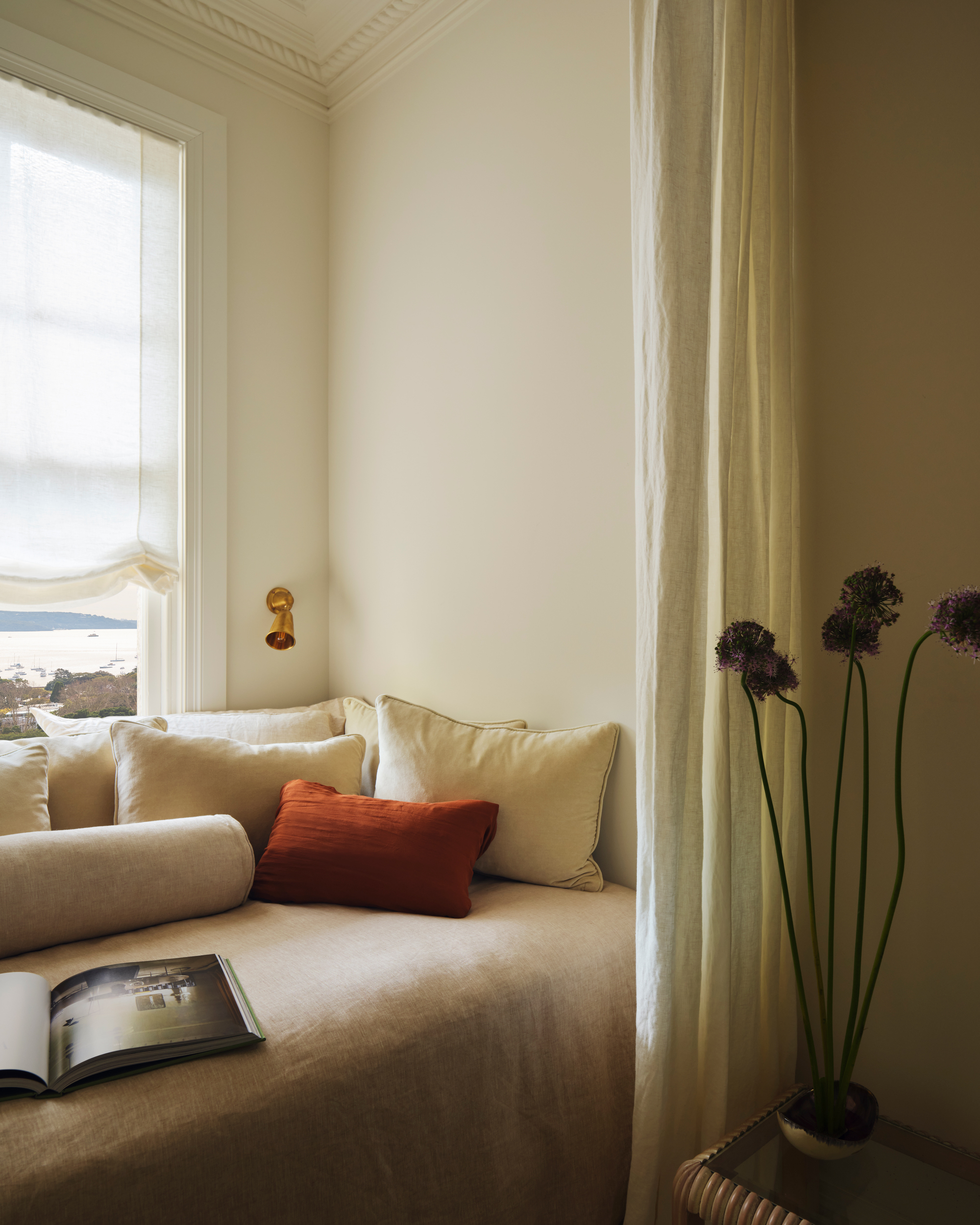 a bed nook by a window with beige bedding and lots of pillows. There is an open book on the bed and a white curtain in front of it.