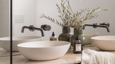 close up of bathroom vanity with two white basins, brushed gunmetal taps, a loewe hand soap, and a vase with olive leaves