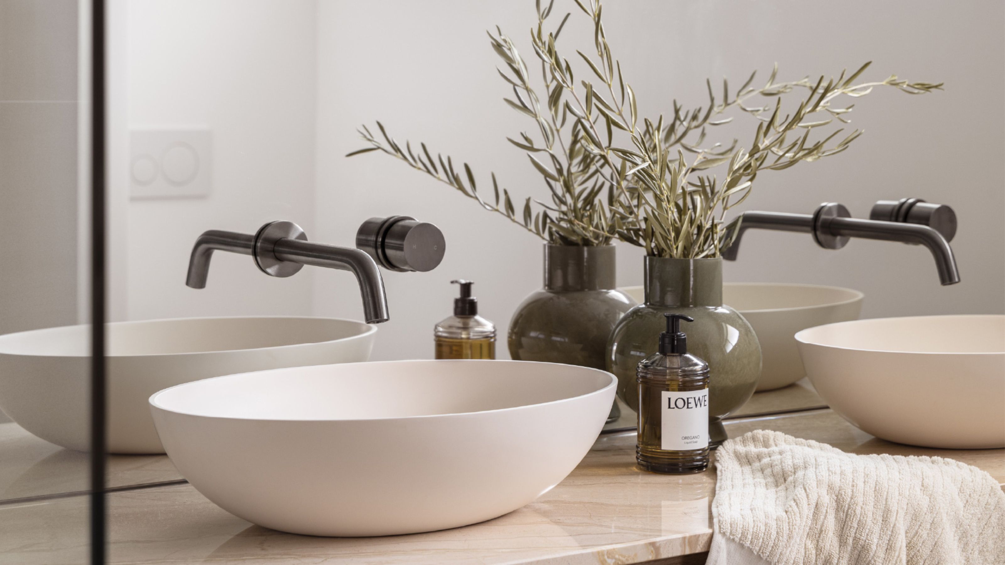 close up of bathroom vanity with two white basins, brushed gunmetal taps, a loewe hand soap, and a vase with olive leaves