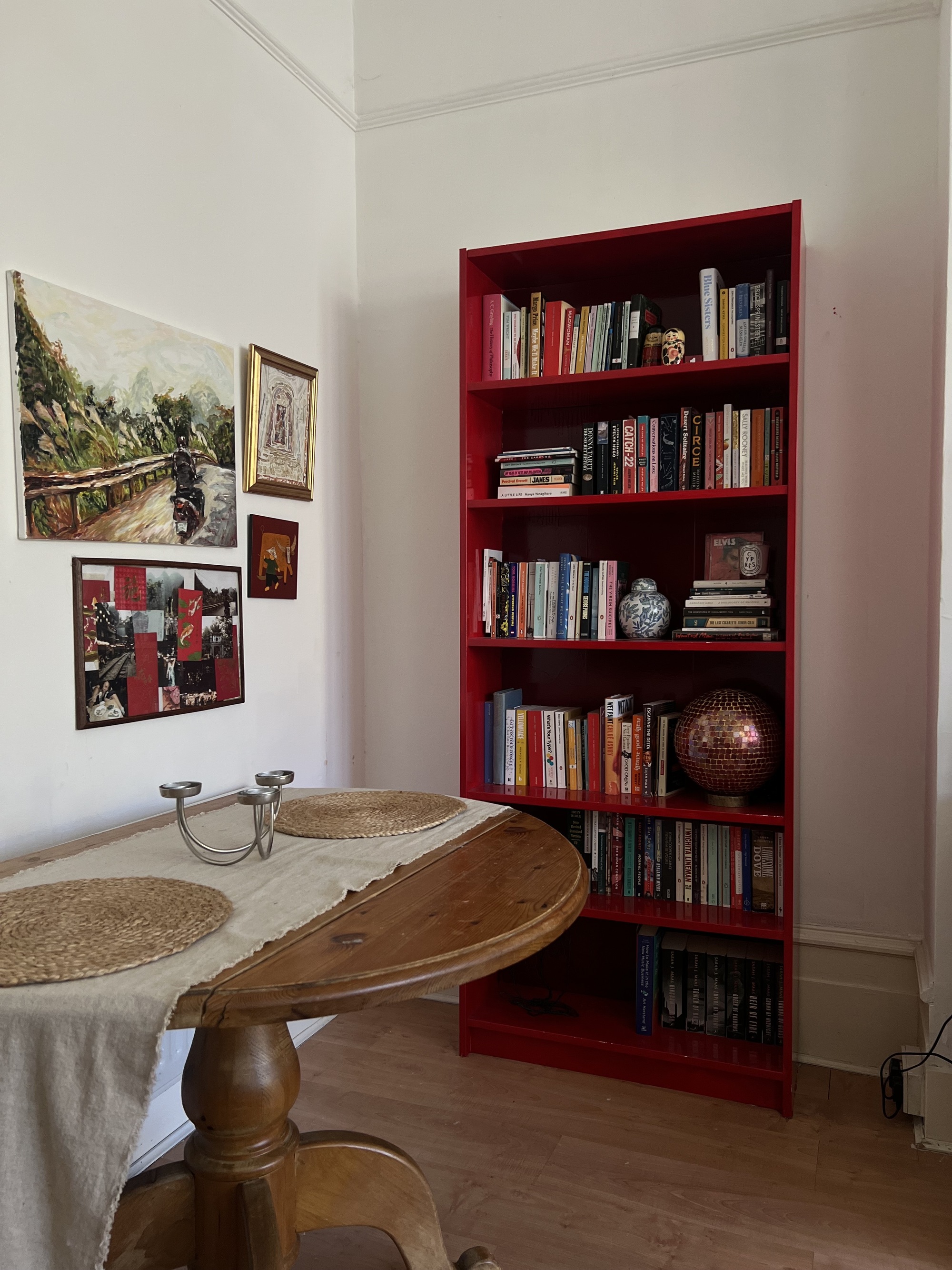 Image of a red bookshelf full of books and decor against a white wall with high ceilings. There is a bistro table in view with a runner, jute placemats, and a silver candle holder. There are four art pieces hanging on the wall.