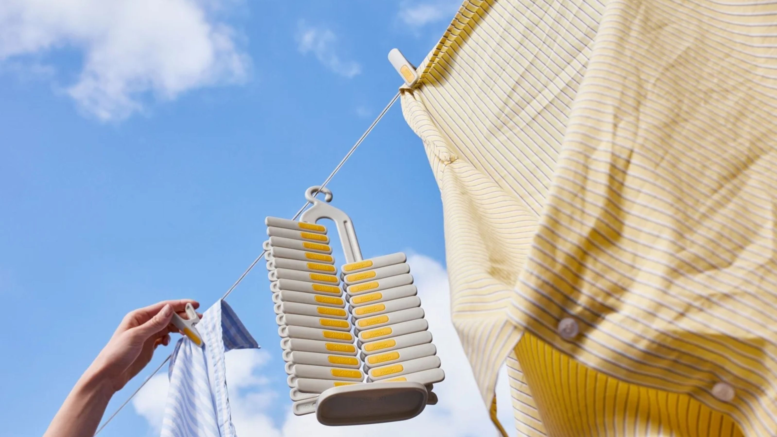 A hand hanging blue and yellow shirts on a washing line with the Joseph Joseph PegStore, with blue skies behind