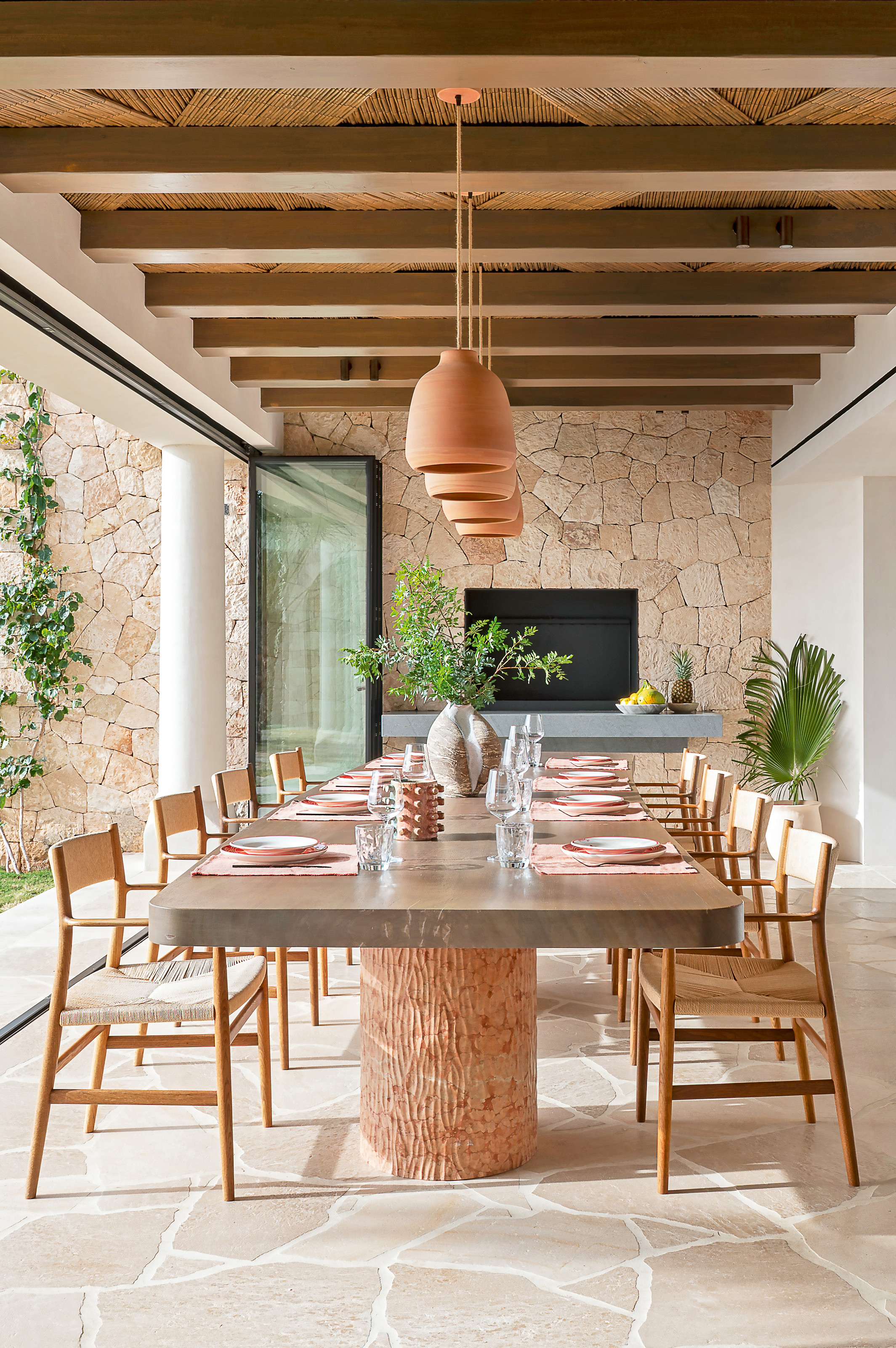 Outdoor dining area with dining table with smooth stone top and carved marble legs, wood and woven seated chairs, terracotta pendant lights overhead