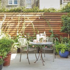 A small, modern garden patio with a table and chairs, along with lots of large, outdoor plants in pots.
