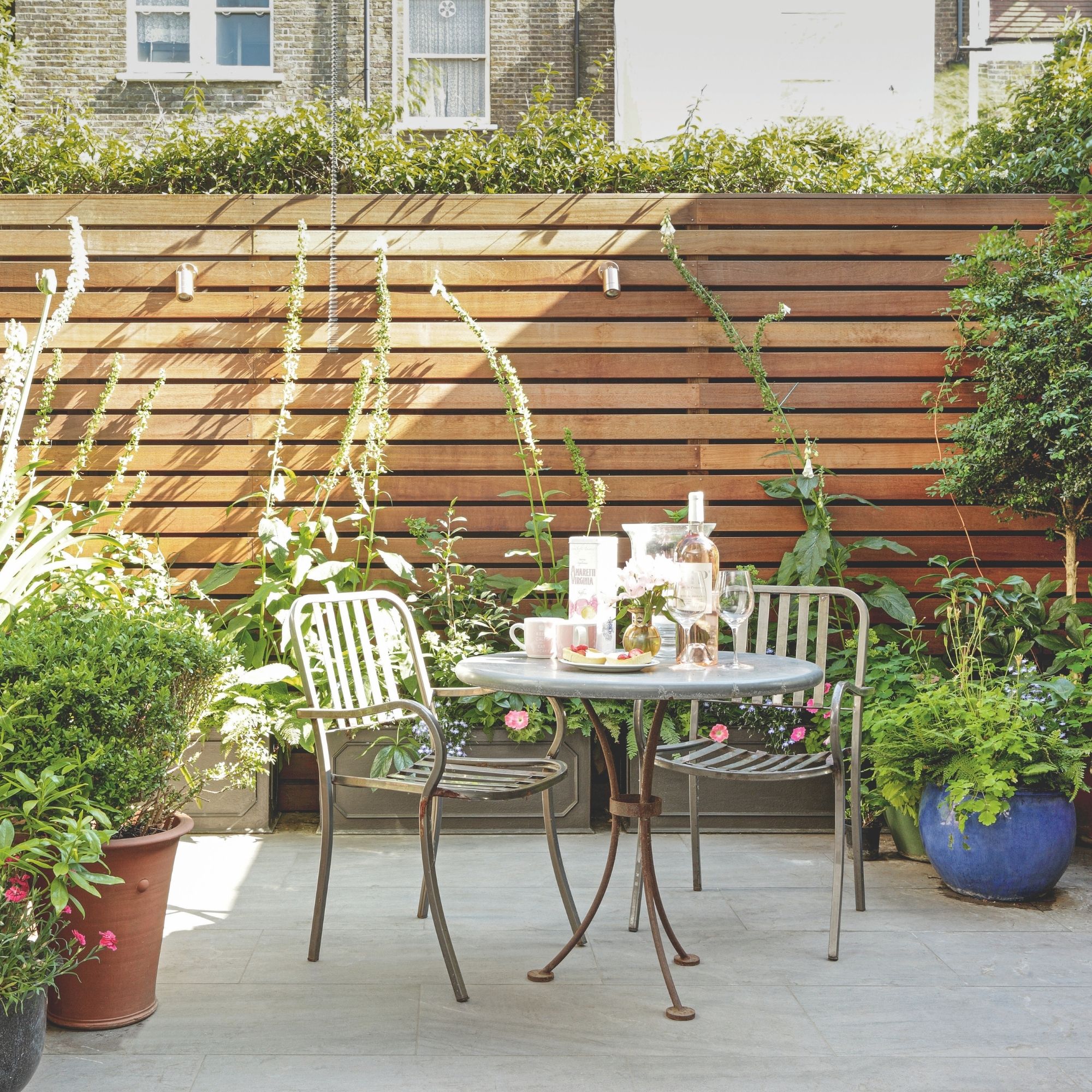 A small, modern garden patio with a table and chairs, along with lots of large, outdoor plants in pots. 