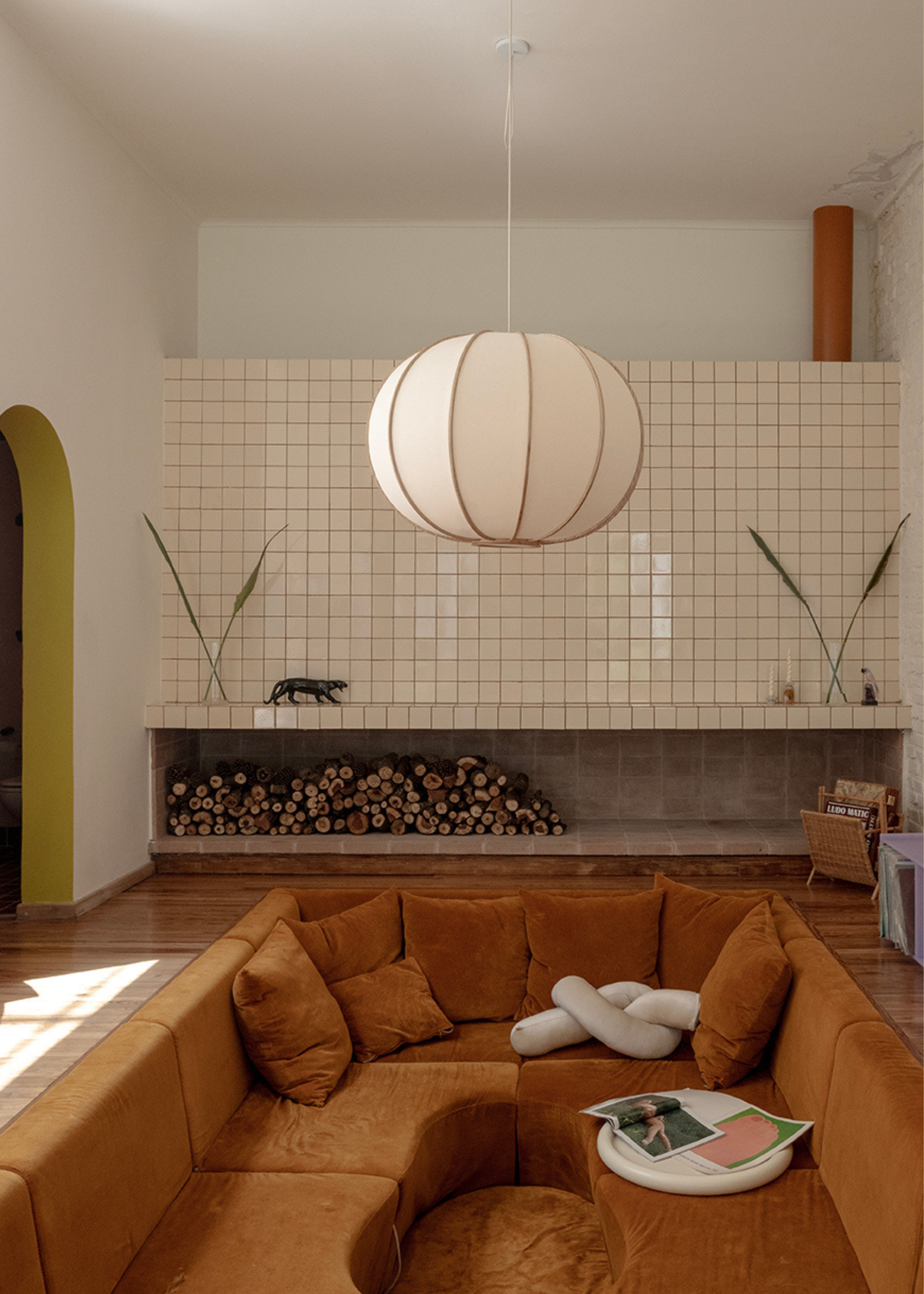 A living room with a tiled wall, a space for logs, a woven magazine rack, an arched doorway with a pop of color, and a deep mustard conversation pit with a bowl