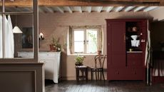 Wide shot of country style laundry and pantry space. There is a freestanding open burgundy larder, wood beam ceiling, bright window and dark hardwood floors