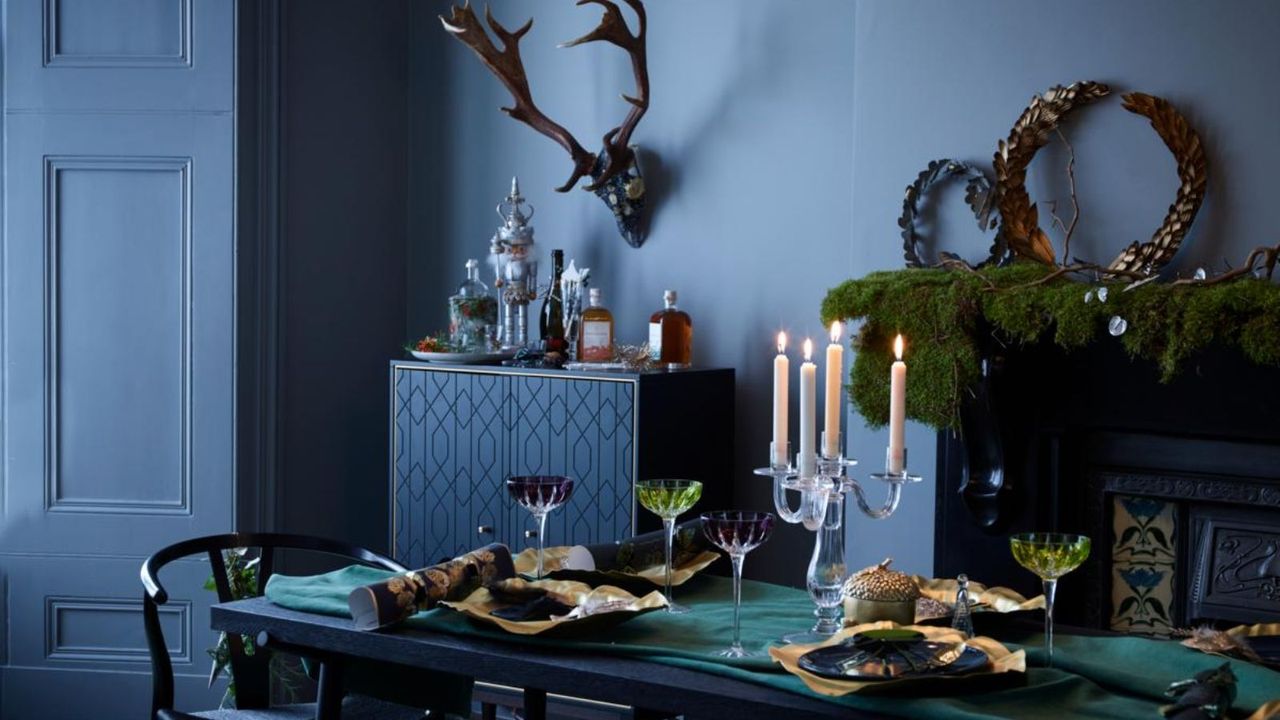 A dining room table set for a Christmas meal, with table settings, blue plates and gold table decorations, a chandelier, stags head on the wall and a mantlepiece with foliage decorations.