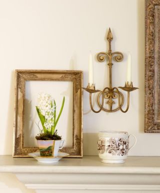 A cream mantel with a brushed gold photo frame, a tea cup with a white flower in it and a an empty floral tea cup, and a gold wall sconce with two candlesticks