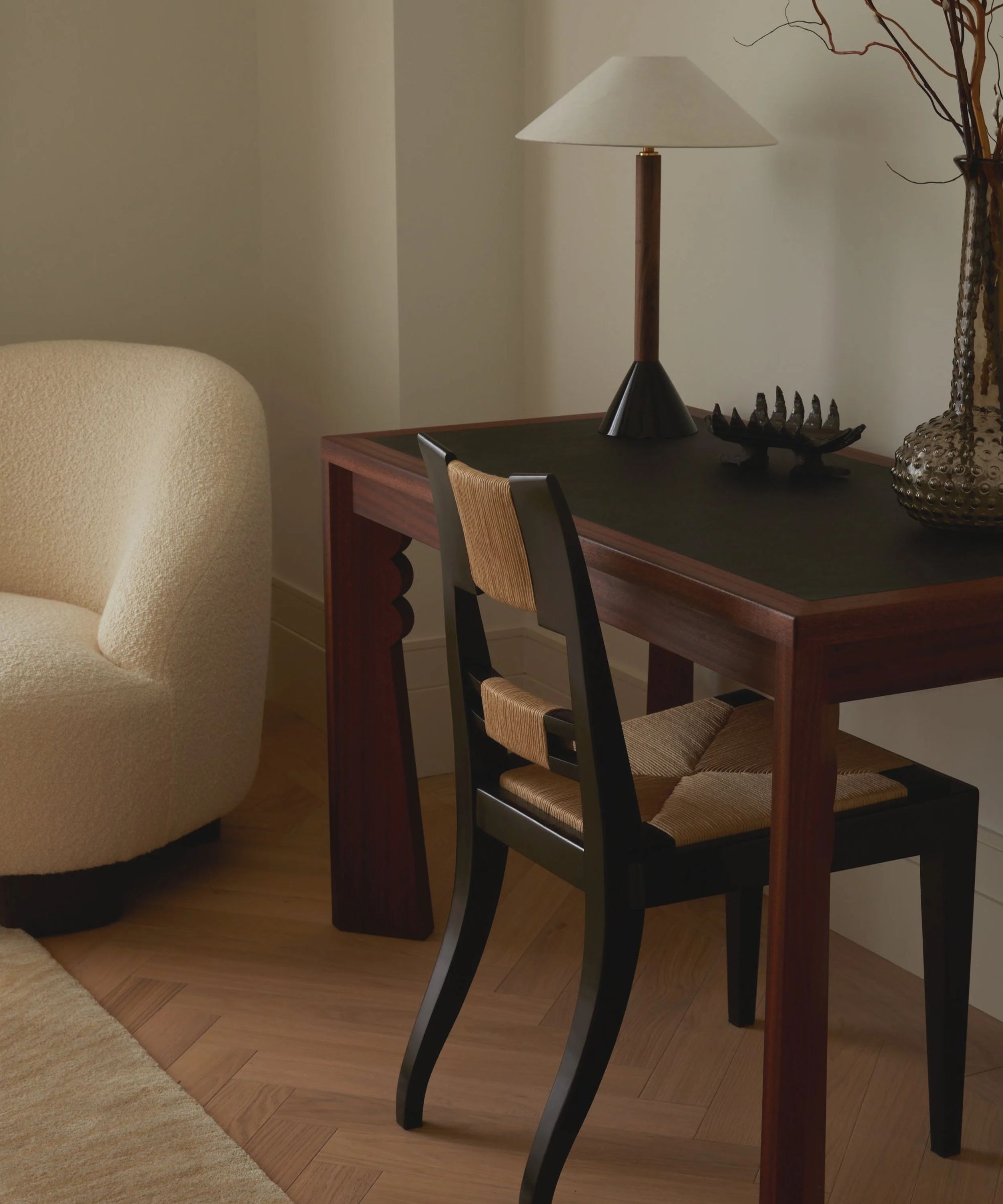 Dark wood desk with dark wood and chair, sculptural table and ornaments, curvacous cream armchair to the left