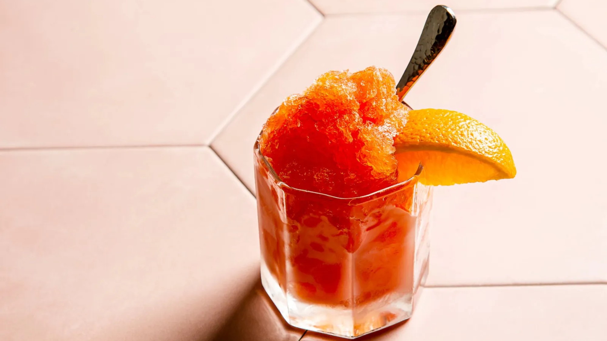3/4 overhead shot of a fluted rocks glass filled with icy garibaldi granita. a spoon and orange wedge included
