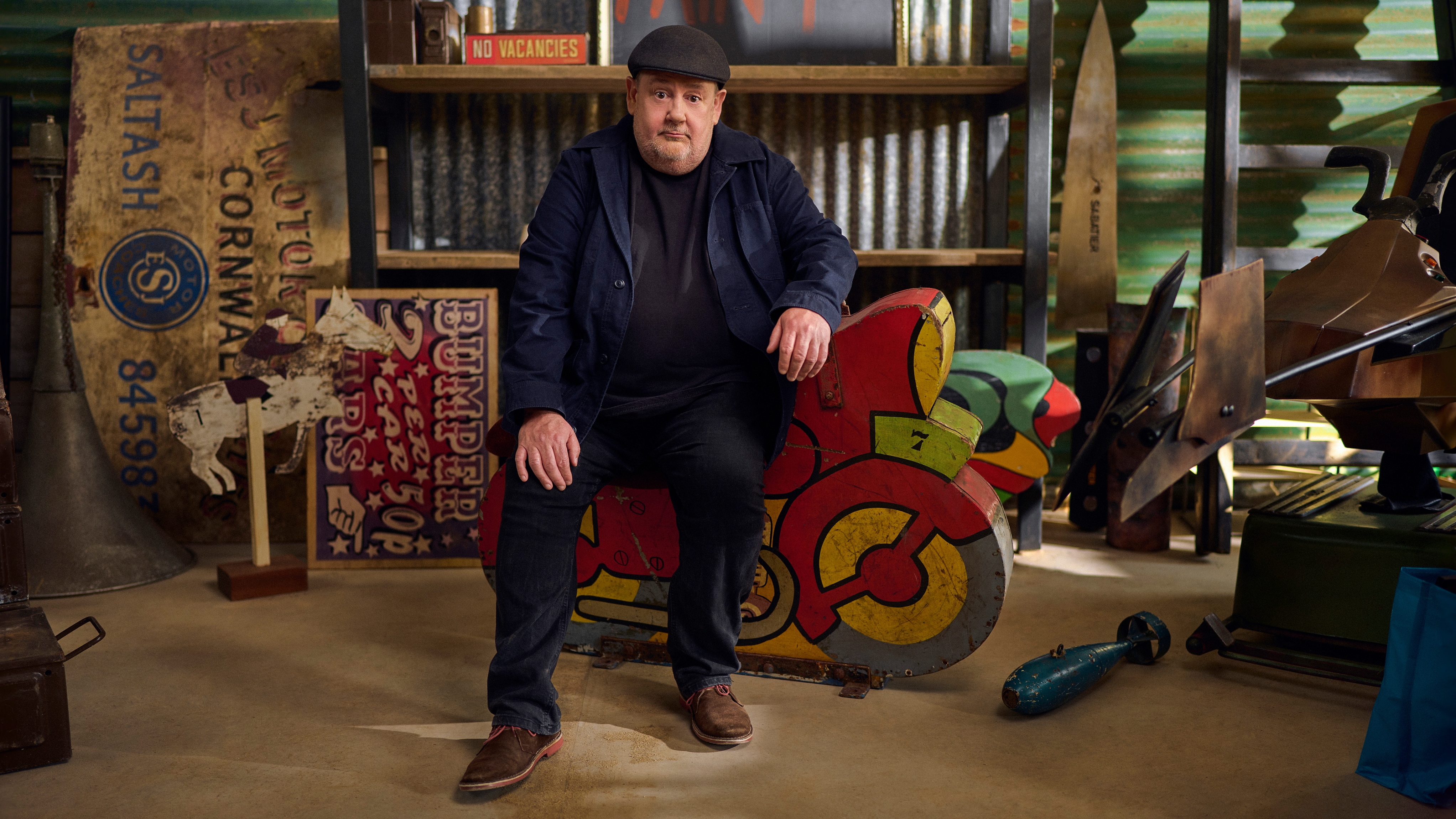 Johnny Vegas sitting in a barn surrounded by antiques