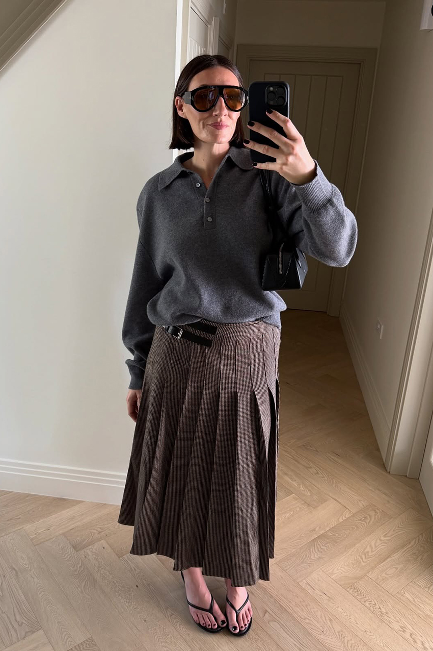 a woman wearing a gray sweater, a kilt plaid skirt, and round black sunglasses