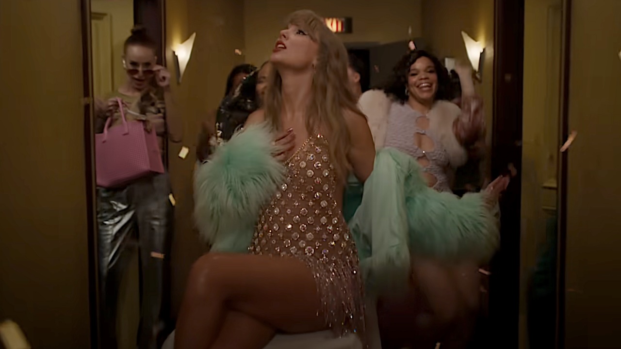 Taylor Swift wearing chain mail and blue fur for The Fate of Ophelia music video. She is singing and sitting on a food cart while dancers dance behind her.