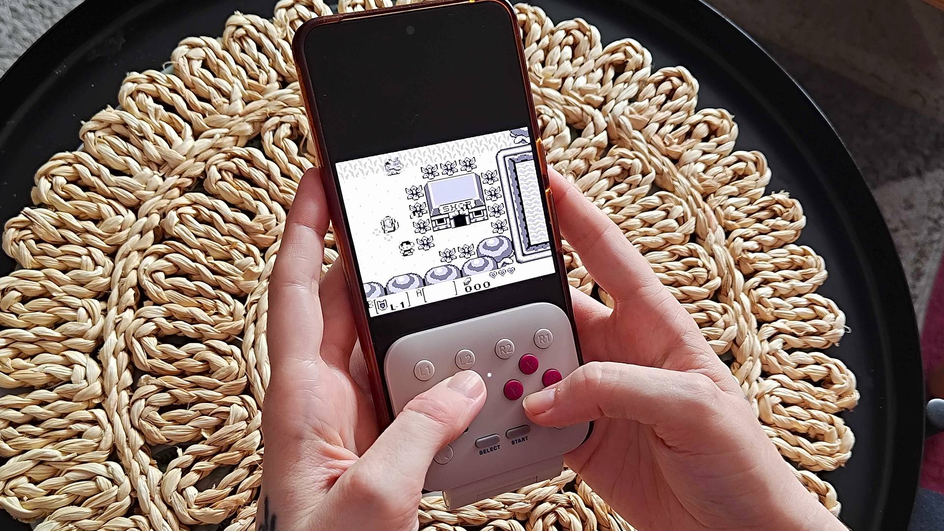 Hands using 8Bitdo FlipPad to play Zelda: Link's Awakening above wicker table cover.