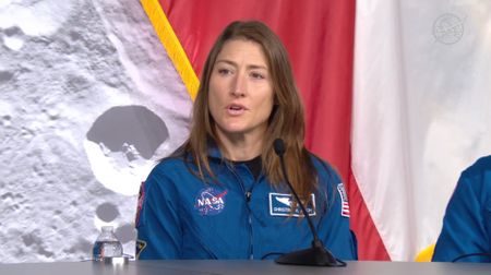 A woman with long dark hair wears a blue flight suit with the circular NASA logo on it and sits behind a desk with a microphone