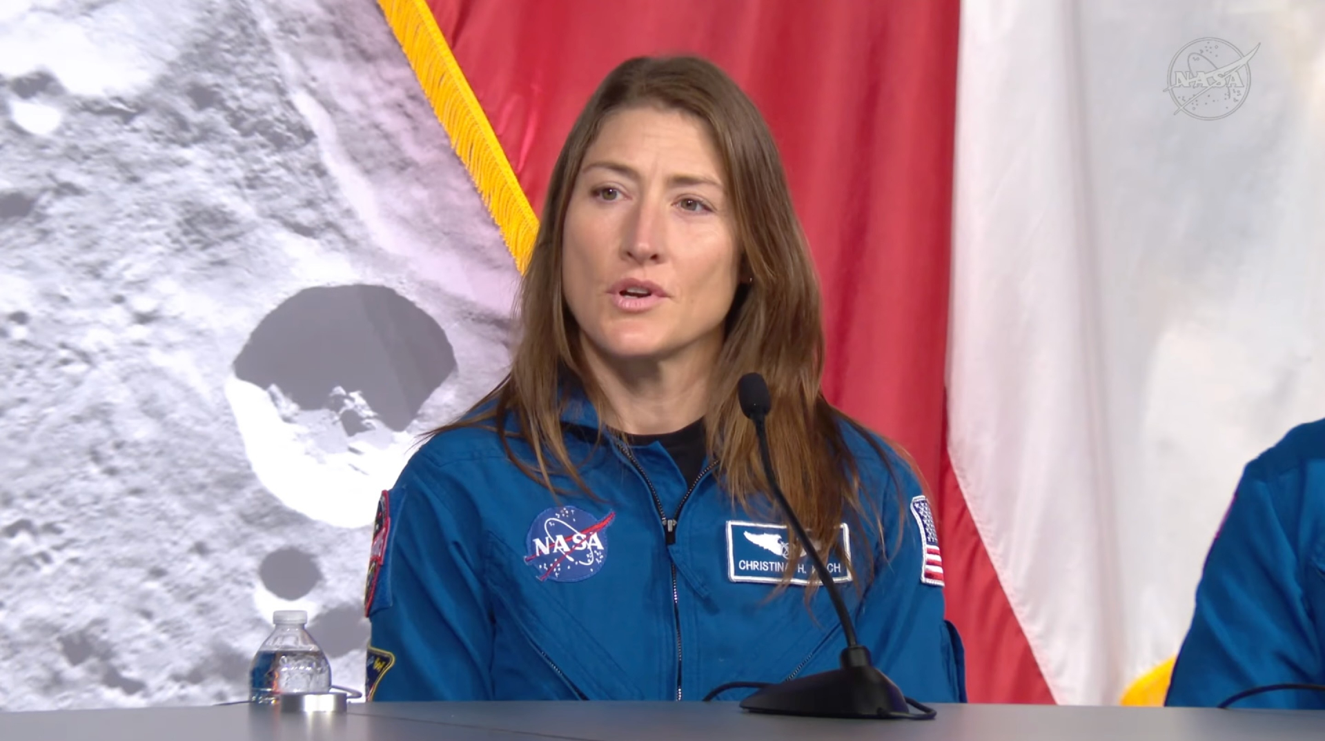 A woman with long dark hair wears a blue flight suit with the circular NASA logo on it and sits behind a desk with a microphone