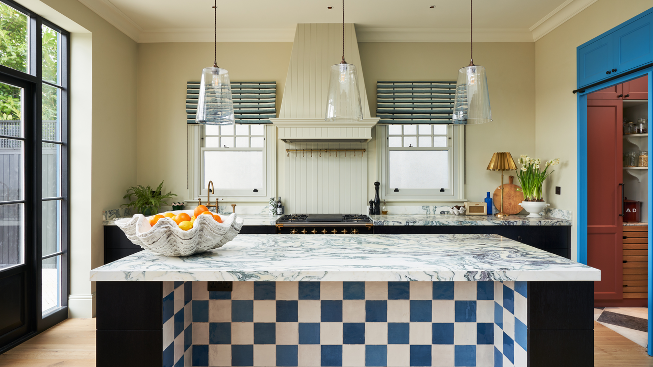 big kitchen with a range cooker and built in extration hood, a blue and white checkered tiled island and marble counters, and bright blue cabinets