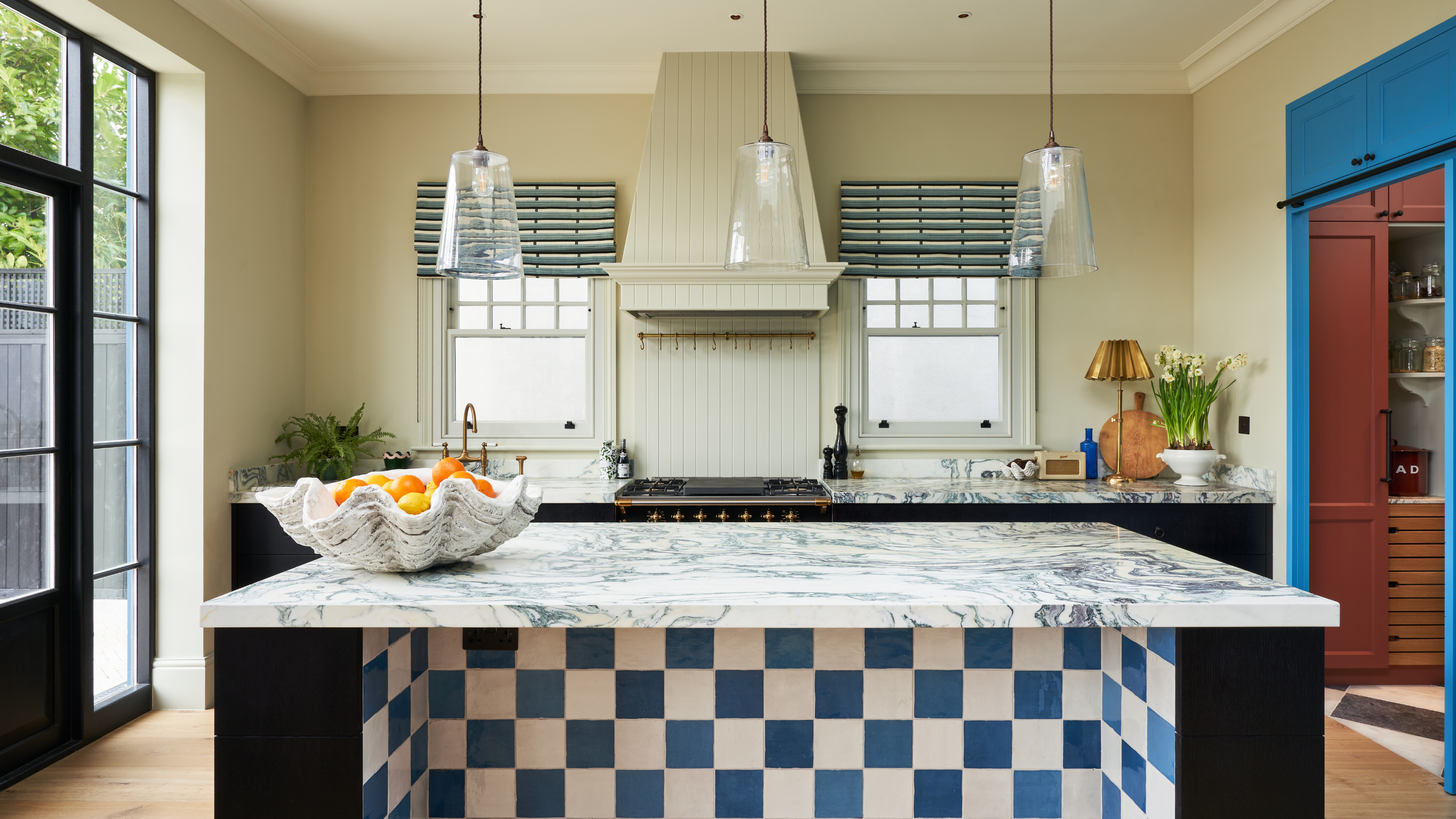 big kitchen with a range cooker and built in extration hood, a blue and white checkered tiled island and marble counters, and bright blue cabinets
