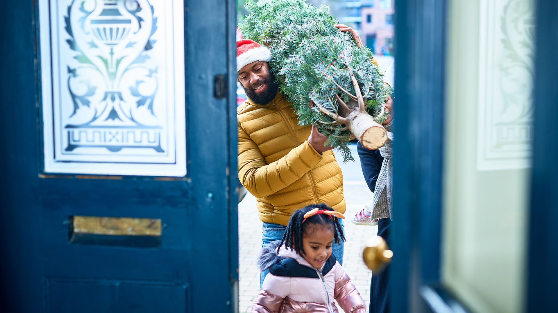 A man walks through a door carrying a Christmas tree with a child walking in the foreground
