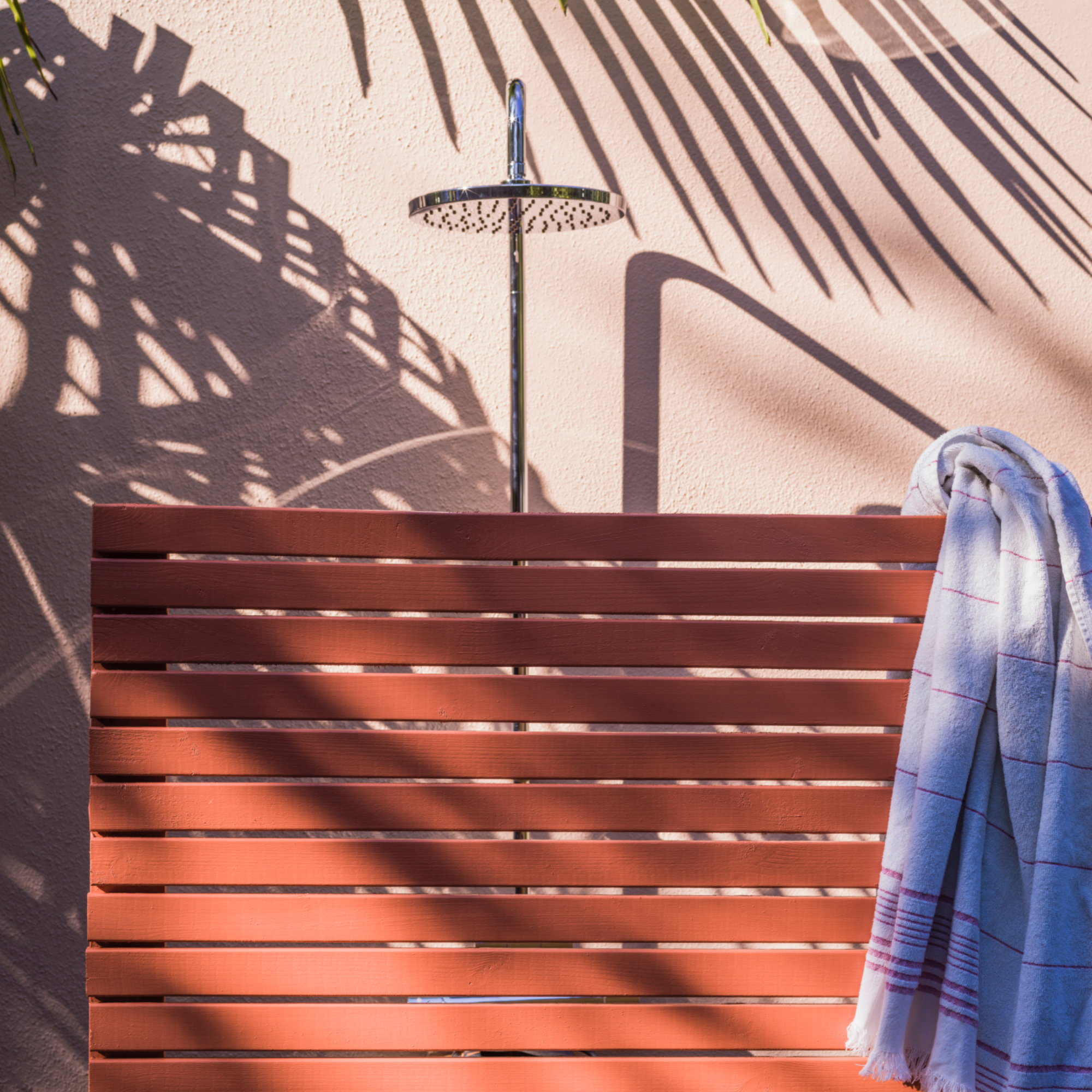 outdoor shower with painted fence screen in terracotta and pale pink wall behind, towel hanging over screen