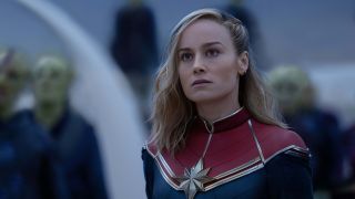 Still from The Marvels (2023) movie. Close up of Captain Marvel (Carol) among green-skinned humanoid aliens known as Skrulls. Captain Marvel has long blonde hair swept over her right shoulder and is wearing a red, blue and gold detailed (star-like emblem in the center of her chest) supersuit looking. She is gazing ahead with a serious look on her face.