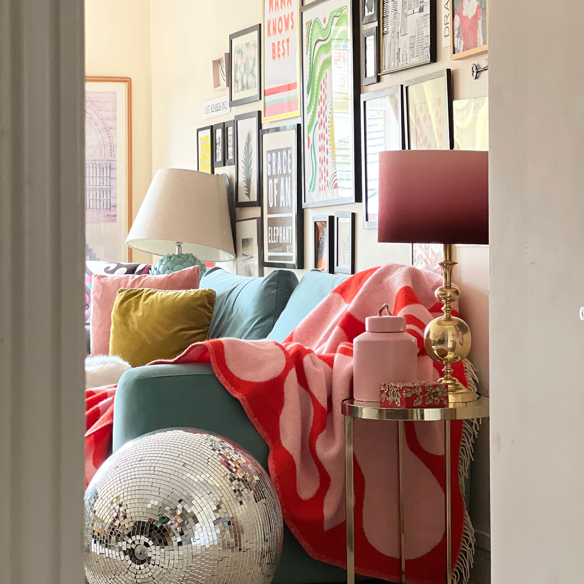View from doorway into living room showing large disco ball on floor beside teal sofa draped with red throw beneath gallery wall of colourful prints
