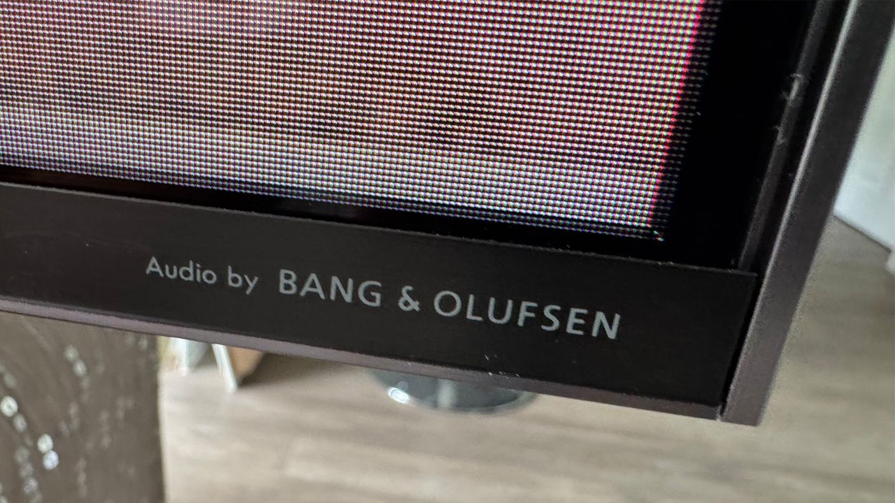 A close-up of the bottom corner of the bezel of the TCL 98C7K 98-inch TV, bearing the legend 'Audio by Bang &amp; Olufsen'.