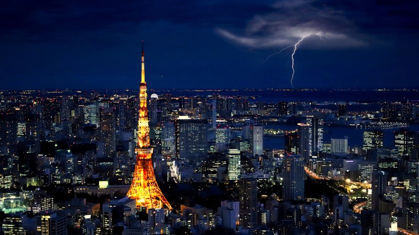 Tokyo skyline at night with a vibrant orange Tokyo Tower illuminated. A bolt of lightning strikes in the distant dark sky, creating a dramatic scene