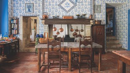French kitchen with antique table and chairs, terracotta tiles, blue tiled walls, large fireplace, copper pans, side table with fruit and veg, steps to the right upstairs