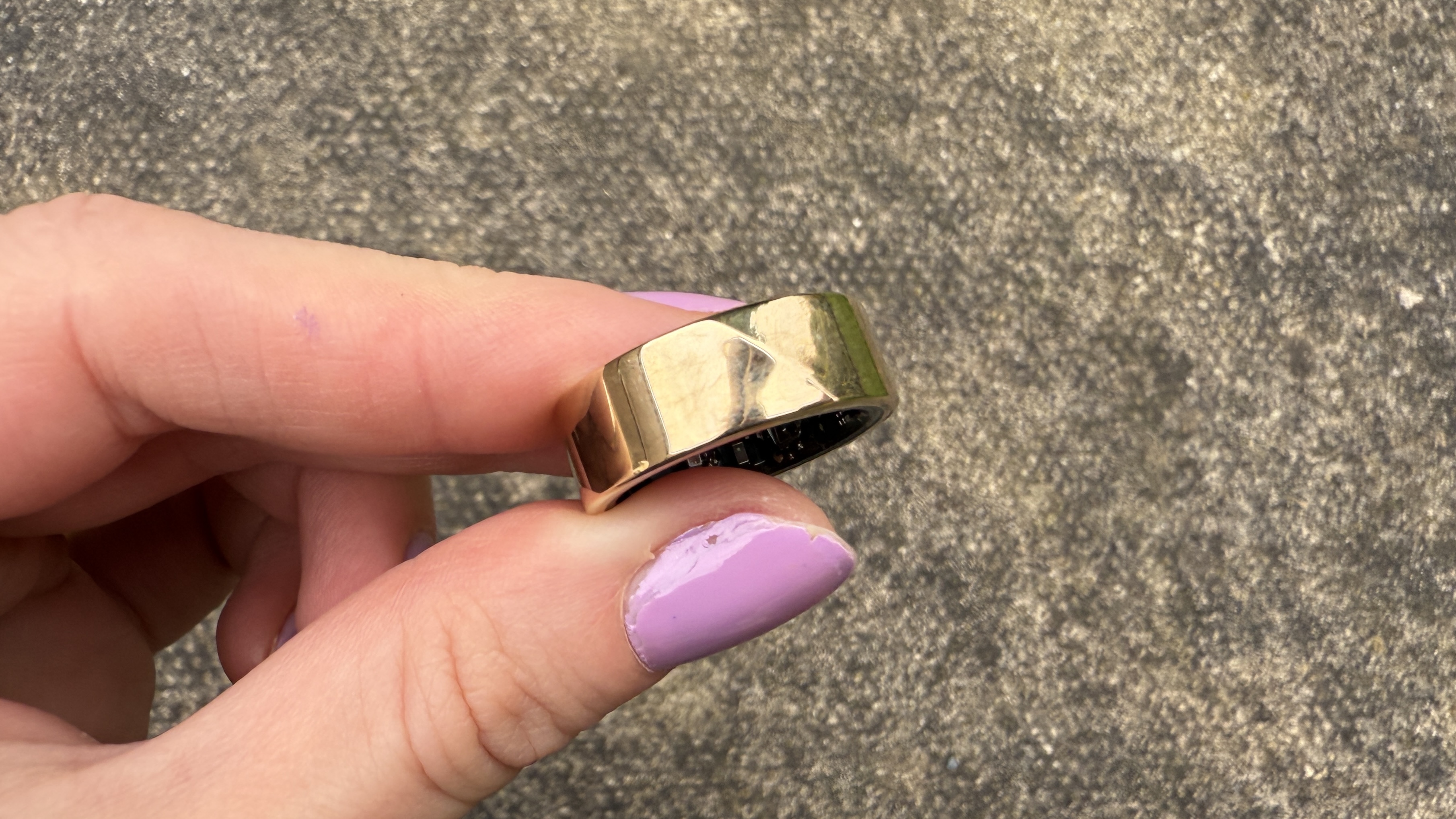 The Luna Ring 2 in sunlit gold against a concrete surface being held between thumb and forefinger