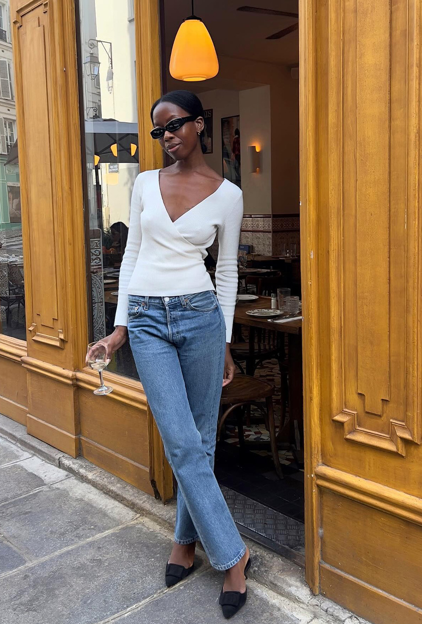 A woman standing in front of a restaurant wearing a white wrap top, cigarette jeans, black kitten heels, and black sunglasses