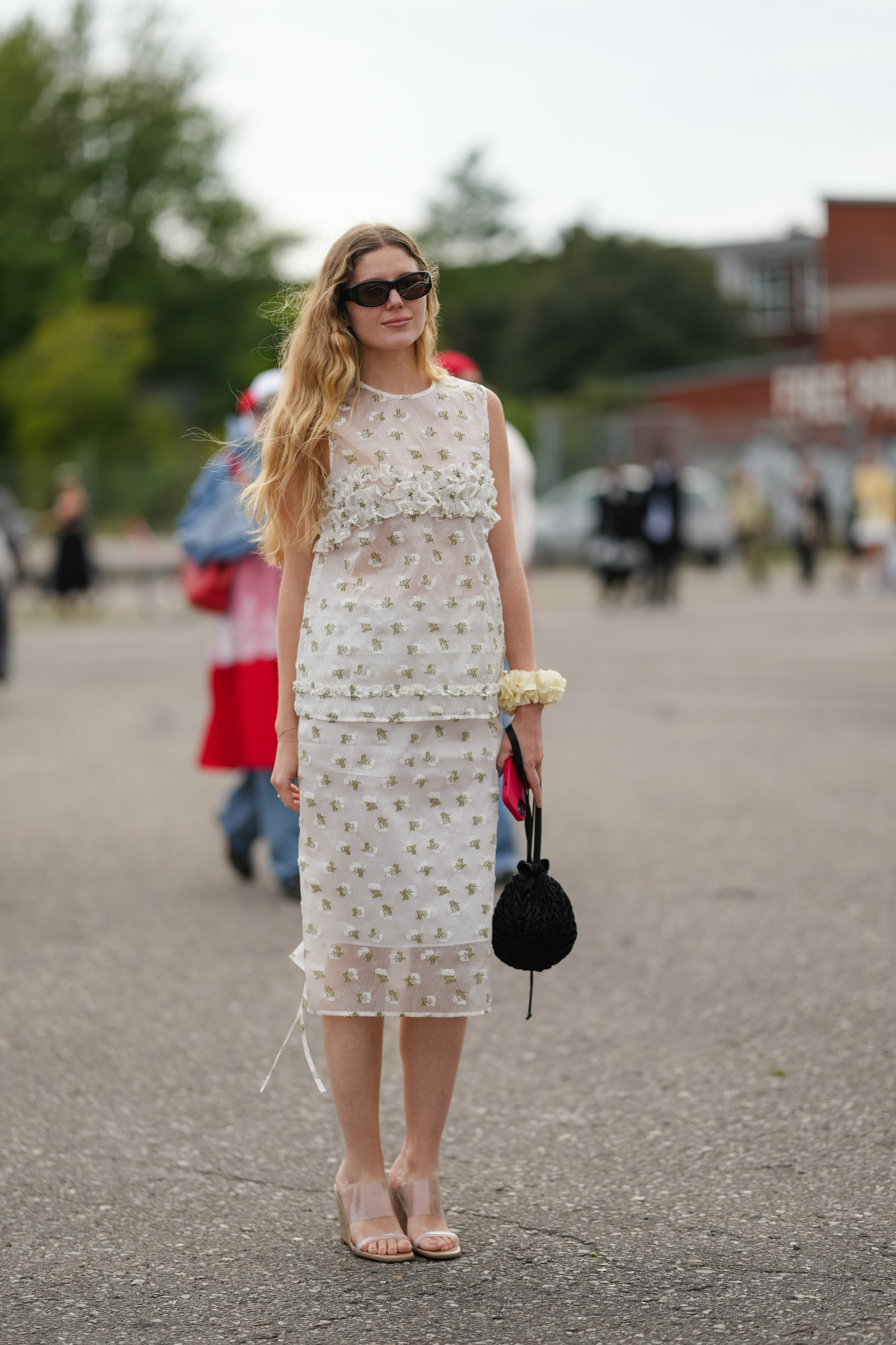 COPENHAGEN, DENMARK - AUGUST 07: A guest wears long wavy blonde hair parted off‑center, black rectangular cat‑eye sunglasses, a sheer ivory tulle sleeveless top scattered with small pale‑gold floral embroidery and trimmed with a horizontal band of ruffled floral appliqu&eacute;s across the chest, a matching sheer tulle midi skirt with the same allover floral embroidery layered over a white slip, an off‑white fabric floral wrist corsage, and a small black crocheted drawstring pouch bag with a wrist strap, finished with clear PVC slide mules with mid‑height block heels and nude footbeds and a clear glossy pedicure, standing while holding the bag, outside Cecilie Bahnsen, during Copenhagen Fashion Week, on August 07, 2025 in Copenhagen, Denmark (Photo by Edward Berthelot/Getty Images)