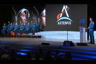 a man in a blue flight suit stands at a lectern on a stage. behind him and to his left is a screen showing the lunar surface, the word "artemis," and a group of people in blue flight suits sitting in chairs.