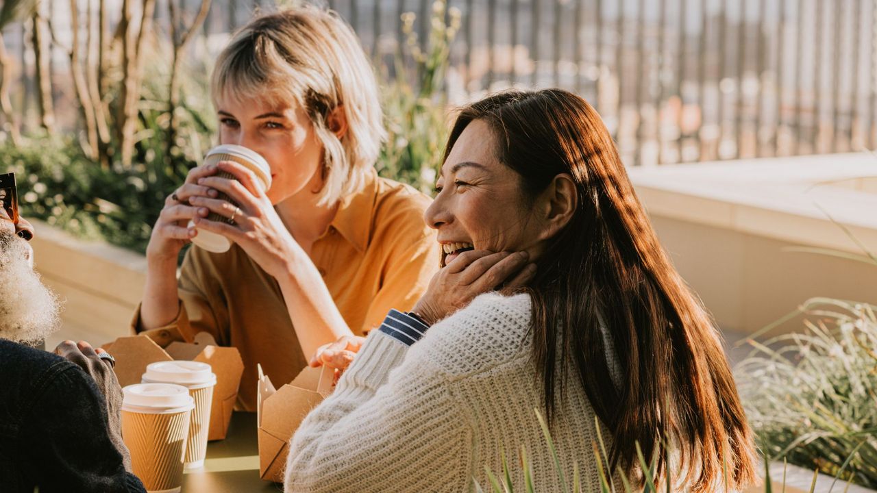 Two women holding and drinking coffee sat round table outside in the sunshine during the morning, representing when to drink coffee